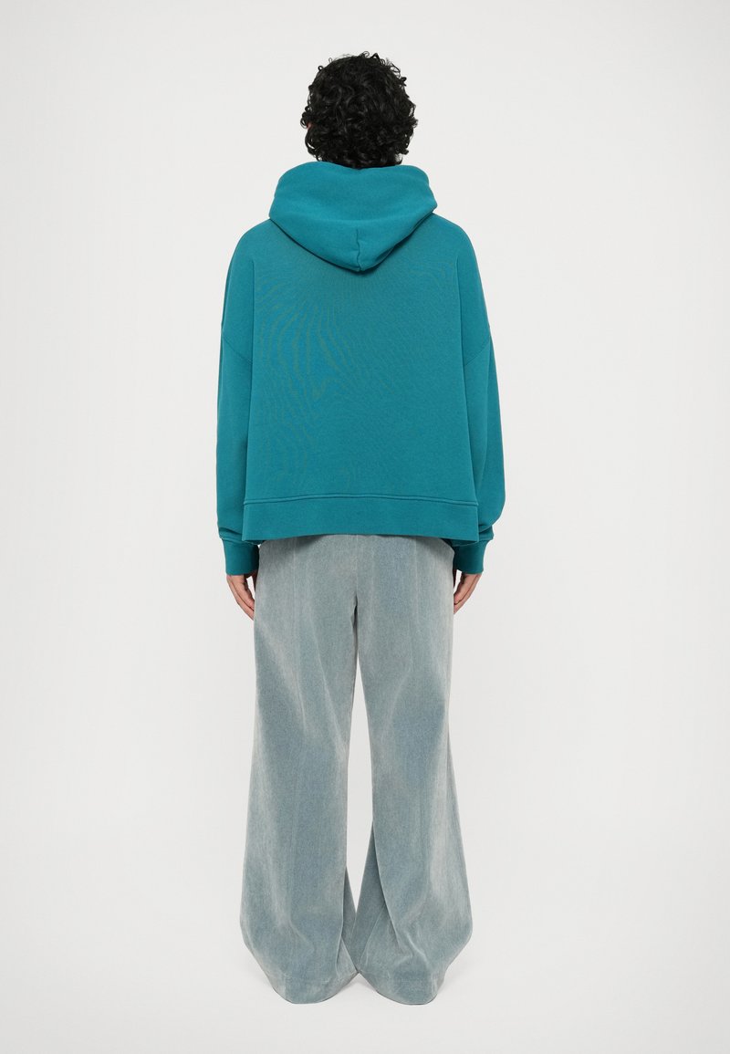 Türkiser oversized Hoodie mit großer Kapuze, der eine subtile Textur aufweist, kombiniert mit hellgrauen, weit geschnittenen Hosen aus einem weichen Stoff.