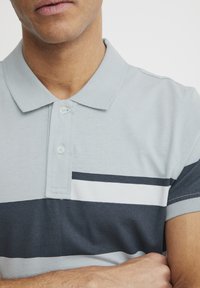 Helehall polo särk kahe nupuga nööbist, millel on tumedhallid ja valged horisontaalsed triibud rinnal ning lühikesed varrukad.