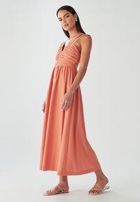 Maxikleit korallivärvilisest kangast, millel on kortsuline ülaosa, õhukesed rihmad ja voolav seelik. Kandmisega tasapinnalised sandaalid. Sile tekstuur.