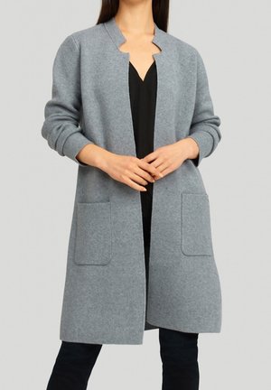 Vrouw die een lange grijze cardigan met open voorkant en opgestikte zakken draagt, over een zwarte top en zwarte broek, staand tegen een effen achtergrond.