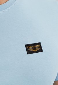 T-shirt en coton bleu clair avec une étiquette rectangulaire noire comportant du texte doré et un design d'ailes, positionnée sur la partie supérieure gauche de la poitrine.