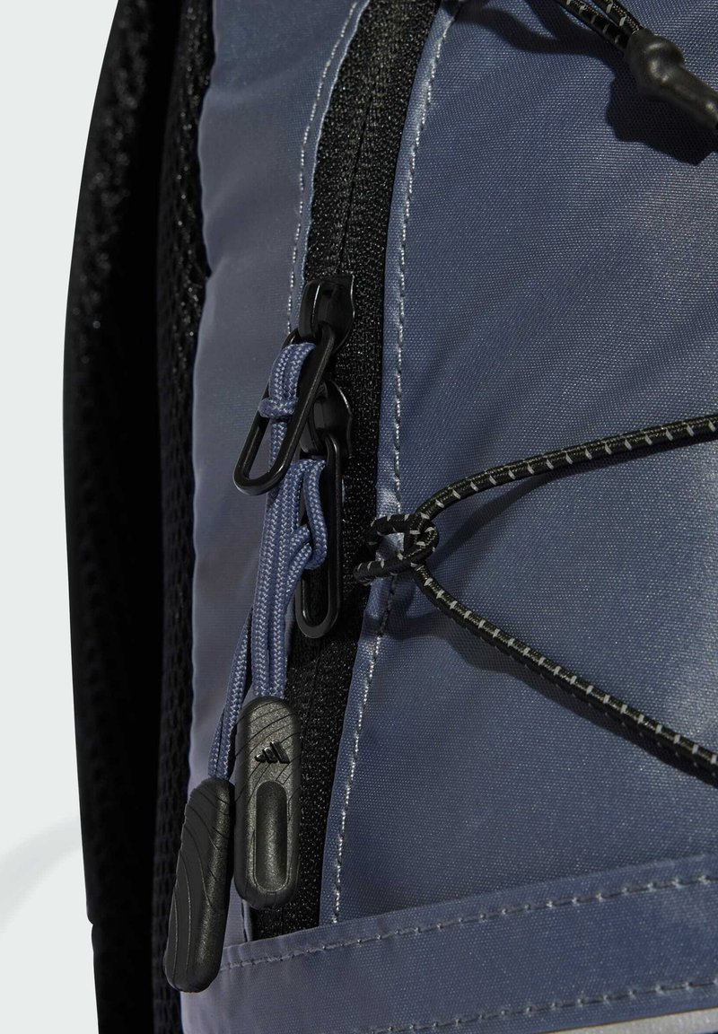 Blauer Stoffrucksack mit schwarzem Netz und Reißverschlüssen; ausgestattet mit doppelten Reißverschlussschiebern mit strukturierten Griffen und einem elastischen Band zur Sicherung von Gegenständen.