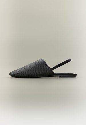 Sort tekstureret flad mule med spids lukket tå og tynd elastisk slingback-rem, sat mod en ensfarvet lys baggrund.