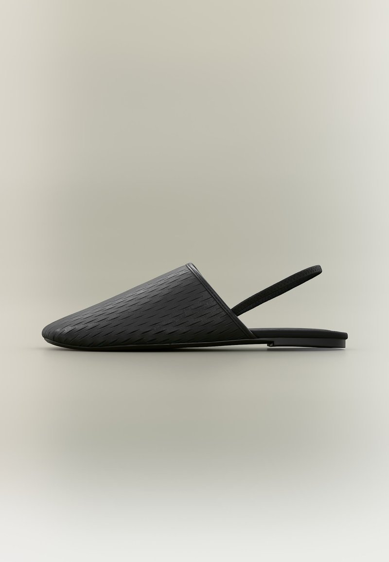 Μαύρο υφασμάτινο, επίπεδο mule με μυτερό κλειστό μπροστινό μέρος και λεπτό ελαστικό λουράκι γύρω από τον αστράγαλο, τοποθετημένο σε απλό ανοιχτό φόντο.