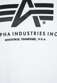 Bijela majica s crnim grafičkim tekstom, na kojoj su istaknuti "ALPHA INDUSTRIES INC" i "KNOXVILLE, TENNESSEE, SAD."