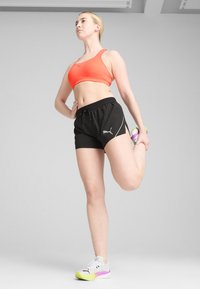 Puma sports bra in bright orange, black shorts with side slits, and white sneakers with purple and green accents. Model stretching.