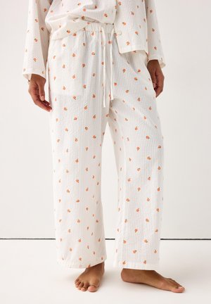 Białe teksturowane spodnie piżamowe z małym pomarańczowym wzorem owocowym, luźne, noszone na boso przez stojącą osobę.