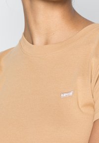 Béžové bavlněné tričko s krátkými rukávy, s malým bílým logem Levi's na levé straně. Hladký žebrovaný vzor na výstřihu.