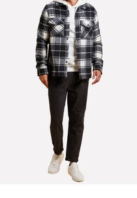 Chemise en flanelle à carreaux noirs et blancs avec deux poches poitrine, superposée sur un sweat à capuche gris, associée à un pantalon foncé et des baskets blanches.