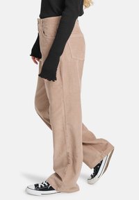 Lichtbruine corduroy broek met wijde pijpen, ribbelstructuur en achterzakken. Gekleed met een zwarte lange mouwen top en zwarte sneakers.