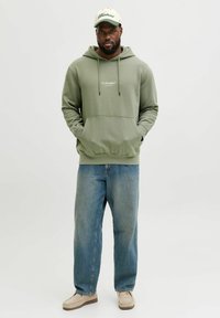 Olivgrön hoodie med framficka och dragsnören, kombinerad med blåa löst sittande jeans och beiga slip-on skor. Kepsen har text.