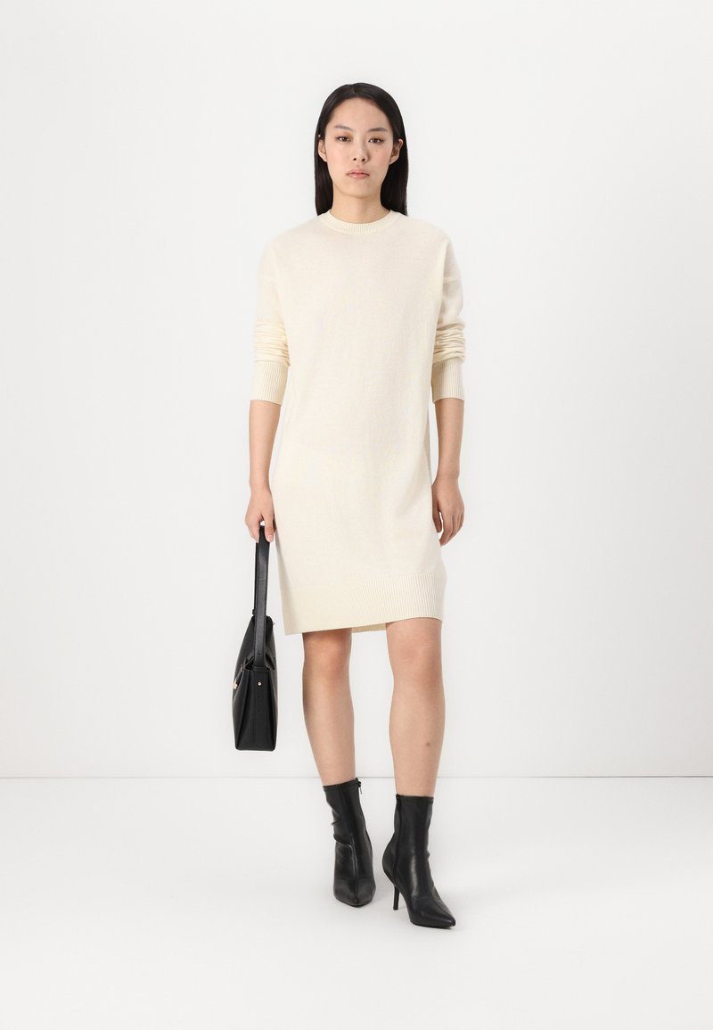 Dzianinowa kremowa sukienka z długimi rękawami i prążkowanymi mankietami, w połączeniu z czarnymi botkami na wysokim obcasie i usztywnioną czarną torebką.