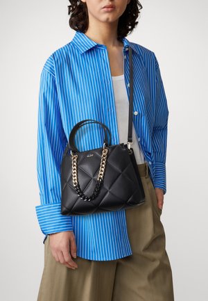 Mujer con camisa azul de rayas y pantalones caqui sostiene un bolso acolchado negro con correa de cadena dorada y negra sobre el hombro.