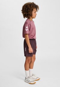 Ungdomssportskläder med en kortärmad rosa topp och mörklila shorts, med dekorativa chevron-accenter och elastisk midja. Vita sportskor.