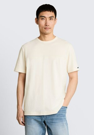 Mężczyzna z krótkimi ciemnymi włosami, ubrany w gładki, jasnobeżowy t-shirt z krótkim rękawem i jasnoniebieskie dżinsy, stojący z jedną ręką w kieszeni.