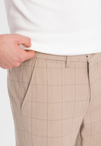 Pantalons à motif carreaux beige en tissu texturé, dotés de poches latérales et d'une fermeture à bouton, présentés avec une main reposant sur la taille.