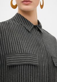 Zwart en grijs gestreept shirt met een kraag, knoopsluiting en twee voorkanten zakken. De stof voelt zacht en iets gestructureerd aan.