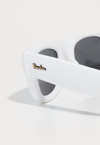 Bijele Ray-Ban sunčane naočale s tamnim staklima, s glatkom teksturom i elegantnim dizajnom. Istaknuti zlatni logo sa strane.