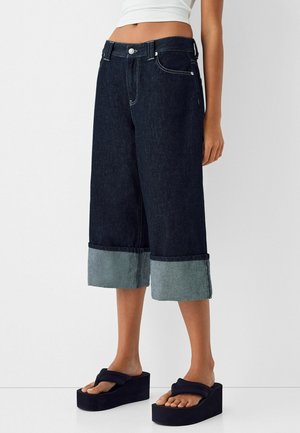 Mørke cropped jeans med vidde ben og brede opklappede manchetter, båret med sorte platformsandaler og hvid crop top.