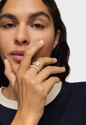 Persona con carnagione media che indossa un anello bianco a forma di orso al dito medio, toccandosi il volto con la mano su uno sfondo chiaro uniforme.