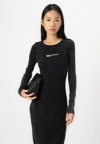 Svart ribbad långärmad klänning med rund halsringning, med "KARL LAGERFELD JEANS"-logotyp. Modellen håller en liten svart handväska.