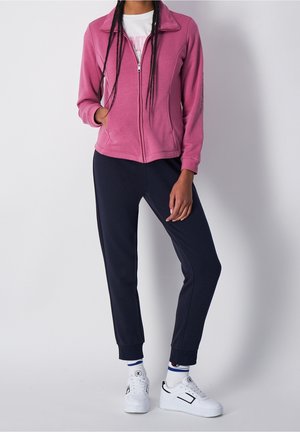 Femme portant une veste zippée rose, un t-shirt blanc avec un motif graphique, un jogging noir, des chaussettes blanches et des baskets blanches, debout devant un fond uni.