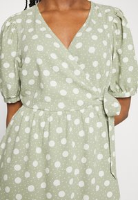 Grön omlottklänning med vita prickar, korta puffärmar och knytning i midjan. Tillverkad av textil med struktur och en flödande kjol.