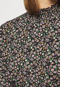 Zwarte bloemenblouse met geplooide halslijn, met een klein bloemmotief in roze, wit, groen en geel op een gestructureerde stof.