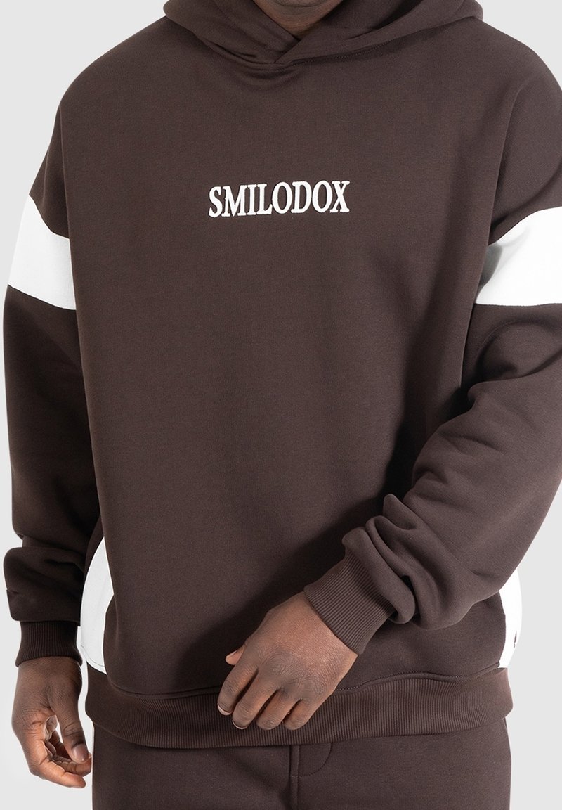 Sweatshirt à capuche marron avec des accents blancs, mettant en avant un logo « SMILODOX » au centre devant. Tissu lisse, poignets et ourlet côtelés.