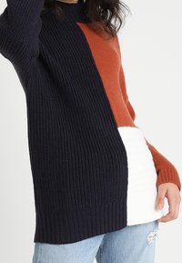 Pull en tricot avec des blocs de couleur bleu marine, rouille et blanc ; texture côtelée ; col rond ; coupe allongée avec une ourlet droit.