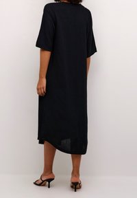 Vestido midi de lino negro con mangas cortas y un corte holgado, que presenta una abertura lateral y una textura suave. Combinado con sandalias de tacón negras.