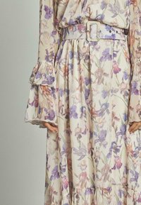 Blumenkleid aus leichtem cremefarbenem Stoff mit lila und pinken Blumenmustern, langen Ärmeln mit Rüschensäumen und einem passenden Gürtel.