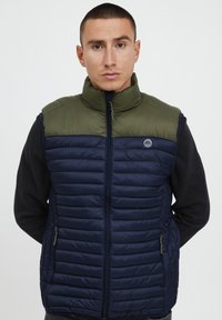 Tamsiai mėlyna siūta liemenė su žaliu apykakle, užtrauktuko uždarymu ir šoninėmis kišenėmis, turinti lygią tekstūrą ir horizontalių siūlių raštą.