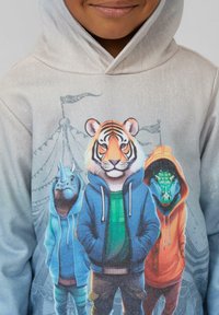 Kind gekleed in een grijze hoodie met een kleurrijke afbeelding van een tijger, neushoorn en krokodil in hoodies, staand voor een circustent.