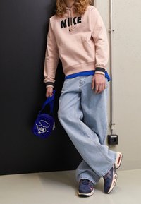 Sudadera rosa claro con logotipo negro de Nike, camiseta azul en capas, vaqueros azules holgados, zapatillas marinas y pequeña bolsa cruzada azul.