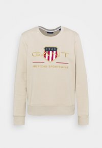 Sweat-shirt beige avec manches retroussées, arborant un grand logo GANT brodé, un design de bouclier, et le texte "American Sportswear" en rouge et or.