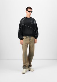 Schwarzer Hoodie mit weißen abstrakten Linien, weite beige Hose und helle Sneakers. Das Modell trägt schwarze Sonnenbrille und steht vor einem weißen Hintergrund.