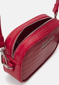 Červená kabelka přes rameno s krokodýlím vzorem a strukturou, vybavená dvěma kapsami na zip a stříbrnými kovovými detaily. Logo vyleptané na boku.
