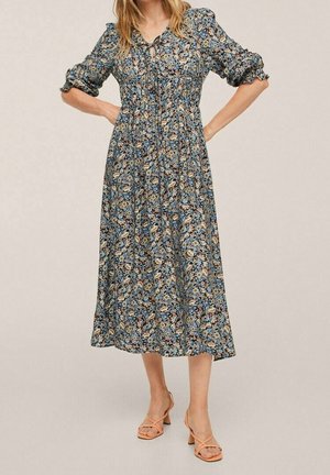 Femme portant une robe midi bleue fleurie avec des manches bouffantes et des poignets à volants, associée à des sandales beiges à talons blocs et lanières.