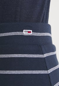 Námořnická ribková tkanina s bílými a šedými horizontálními pruhy. Obsahuje malé vyšívané logo v bílé, červené a modré barvě poblíž pásku.