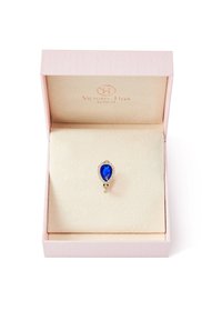 Anello placcato oro con una pietra a forma di pera blu profondo, circondata da cristalli trasparenti, presentato in una scatola regalo rosa pallido con un coperchio testurizzato.