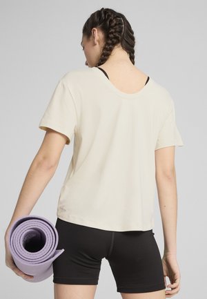 Camiseta de algodón crema con mangas cortas y escote redondeado, combinada con pantalones cortos negros. Porta en su mano un mat de yoga morado enrollado.