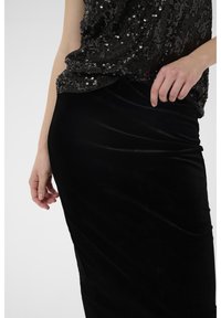 Juodas blizgančiais papuoštas viršus su laisvu siluetu, derinamas su aptakiu, prigludusiu juodu aksomu sijonu. Matoma lygi tekstūra ir blizgantys akcentai.