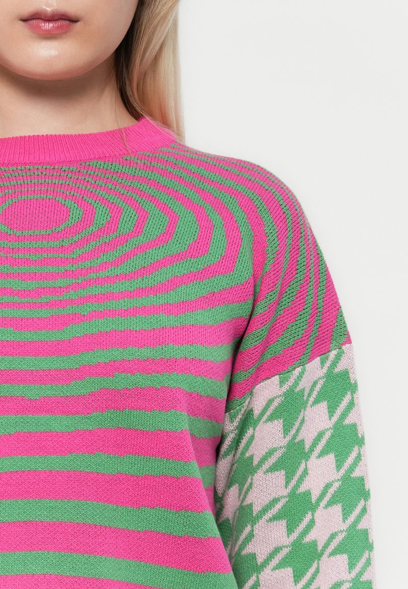 Sweter z dzianiny w poziome różowe i zielone pasy; jedno rękaw ma wzór w szkocką kratę w zielonym i białym kolorze; ściągacz przy dekolcie.