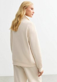 Felpa color beige chiaro con colletto alto, vestibilità comoda e polsini a costine. Texture morbida, design minimalista, abbinata a pantaloni coordinati.