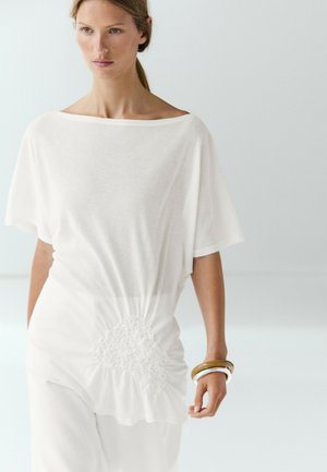 Femme portant un haut blanc ample avec un détail froncé à la taille et un pantalon blanc large, accessoirisée avec des bracelets épais au poignet.