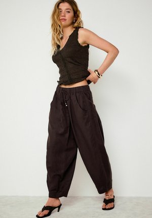 Kobieta o długich blond włosach, ubrana w czarny, bezrękawnik o fakturowanej strukturze, luźne czarne spodnie z troczkiem oraz czarne sandały na obcasie, pozująca na tle gładkiej ściany.