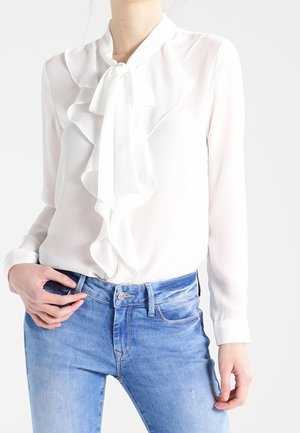 Kvinde iført en hvid bluse med lange ærmer med flæser foran og sløjfe, kombineret med lyseblå denimjeans.