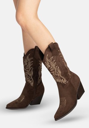 Ben iført brune ruskind cowboystøvler til midt på læggen med beige broderede mønstre og stablede hæle, vist mod en ensartet lys baggrund.