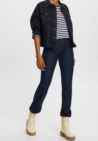 Giacca di jeans blu scuro con due tasche sul petto. Camicia a righe bianche e blu navy, jeans scuri a vita media e stivaletti beige con suola spessa.